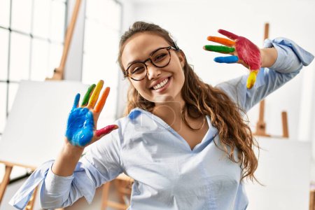 Photo for Young beautiful hispanic woman artist smiling confident showing painted hands at art studio - Royalty Free Image