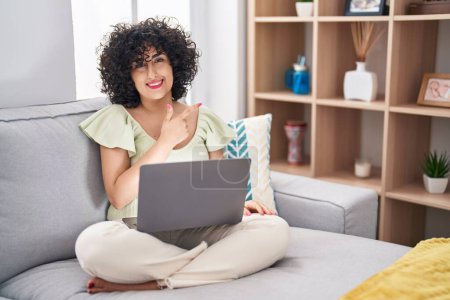 Photo for Young brunette woman with curly hair using laptop sitting on the sofa at home cheerful with a smile of face pointing with hand and finger up to the side with happy and natural expression on face - Royalty Free Image
