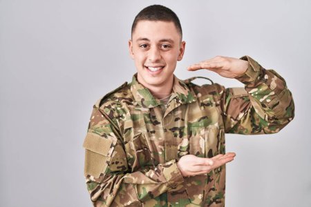 Téléchargez les photos : Jeune homme portant uniforme de camouflage de l'armée geste avec les mains montrant grand et grand signe de taille, symbole de mesure. souriant en regardant la caméra. concept de mesure. - en image libre de droit