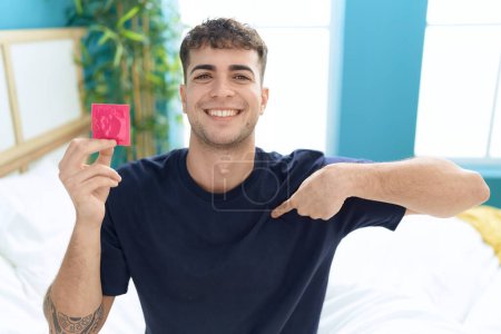 Téléchargez les photos : Jeune homme hispanique assis sur le lit tenant préservatif pointant du doigt un sourire heureux et fier - en image libre de droit