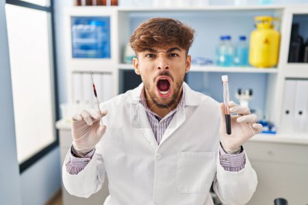 Téléchargez les photos : Homme arabe avec barbe travaillant au laboratoire scientifique tenant échantillon de sang en colère et fou hurlant frustré et furieux, criant de colère. rage et concept agressif. - en image libre de droit