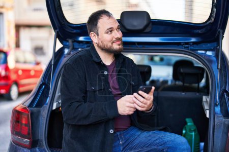 Foto de Hombre hispano joven usando teléfono inteligente sentado en el maletero del coche en la calle - Imagen libre de derechos