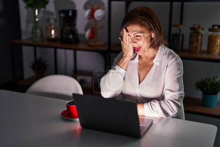 Foto de Mujer hispana de mediana edad usando portátil en casa por la noche cubriendo un ojo con la mano, sonrisa confiada en la cara y emoción sorpresa. - Imagen libre de derechos