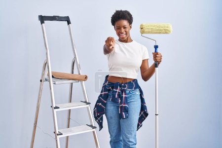 Foto de Mujer afroamericana sosteniendo pintor de rodillos señalándote a ti y a la cámara con los dedos, sonriendo positiva y alegre - Imagen libre de derechos