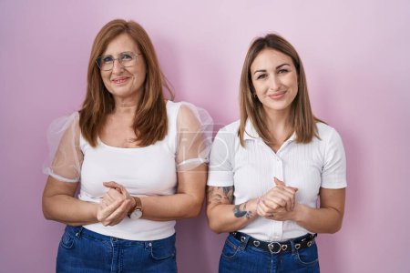 Foto de Madre e hija hispanas llevando una camiseta blanca casual sobre fondo rosa con las manos juntas y los dedos cruzados sonriendo relajadas y alegres. éxito y optimismo - Imagen libre de derechos