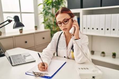 Téléchargez les photos : Young caucasian woman doctor talking on telephone writing on document at clinic - en image libre de droit