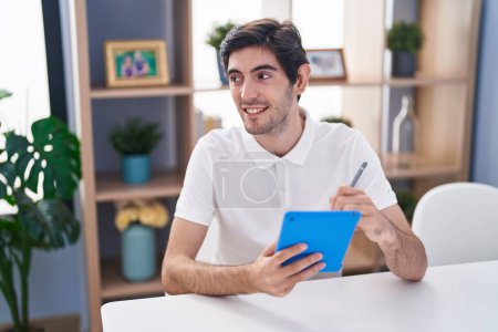 Photo for Young hispanic man writing on touchpad sitting on table at home - Royalty Free Image