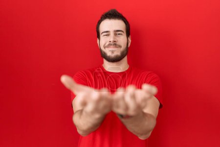 Téléchargez les photos : Jeune homme hispanique portant un t-shirt rouge décontracté souriant avec les mains paumes ensemble recevant ou donnant geste. maintien et protection - en image libre de droit