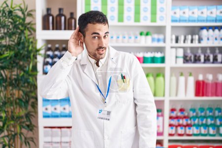 Photo for Handsome hispanic man working at pharmacy drugstore smiling with hand over ear listening an hearing to rumor or gossip. deafness concept. - Royalty Free Image