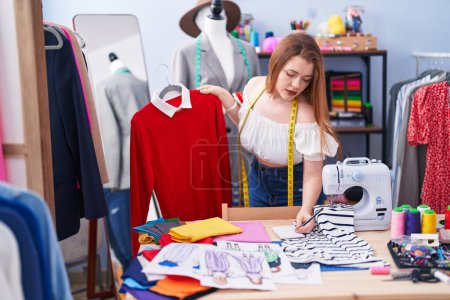 Téléchargez les photos : Jeune femme rousse tailleur tenant t-shirt écriture sur ordinateur portable à l'usine de vêtements - en image libre de droit