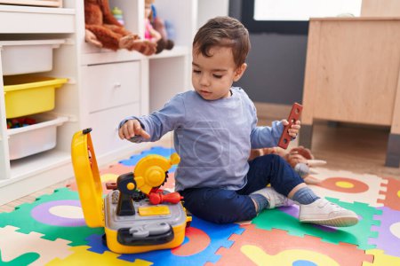Téléchargez les photos : Adorable garçon hispanique jouant avec des outils jouet assis sur le sol à la maternelle - en image libre de droit