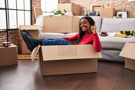 Photo for Young african american with braids moving to a new home inside of a cardboard box surprised with an idea or question pointing finger with happy face, number one - Royalty Free Image