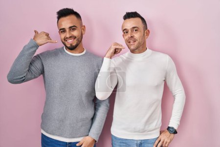 Téléchargez les photos : Couple homosexuel debout sur fond rose souriant faire geste de téléphone avec la main et les doigts comme parler au téléphone. communiquer les concepts. - en image libre de droit