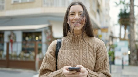 Téléchargez les photos : Jeune belle femme hispanique utilisant smartphone souriant à la terrasse du café - en image libre de droit