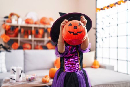 Foto de Adorable chica hispana teniendo fiesta de halloween sosteniendo cesta de calabaza en la cara en casa - Imagen libre de derechos