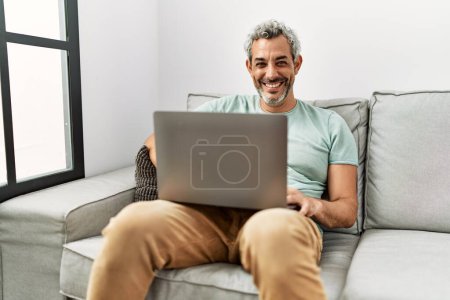 Photo for Middle age grey-haired man using laptop sitting on sofa at home - Royalty Free Image