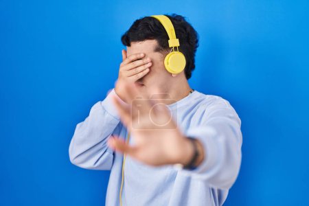 Photo for Non binary person listening to music using headphones covering eyes with hands and doing stop gesture with sad and fear expression. embarrassed and negative concept. - Royalty Free Image
