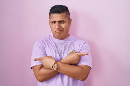 Téléchargez les photos : Jeune homme hispanique debout sur fond rose pointant des deux côtés avec les doigts, direction différente en désaccord - en image libre de droit