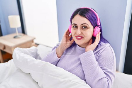 Téléchargez les photos : Jeune belle femme plus la taille écouter de la musique assis sur le lit dans la chambre - en image libre de droit