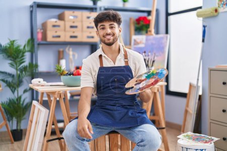Photo for Arab man with beard painter sitting at art studio holding palette looking positive and happy standing and smiling with a confident smile showing teeth - Royalty Free Image