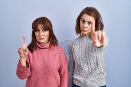 Téléchargez les photos : Mère et fille debout sur fond bleu pointant le doigt vers le haut et l'expression en colère, ne montrant aucun geste - en image libre de droit