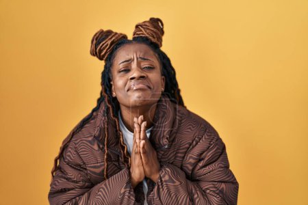 Téléchargez les photos : Femme africaine avec les cheveux tressés debout sur fond jaune mendier et prier avec les mains ainsi que l'expression de l'espoir sur le visage très émotionnel et inquiet. mendier. - en image libre de droit