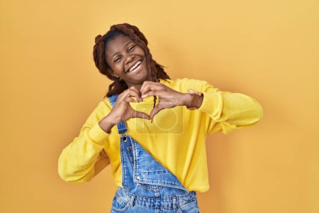 Téléchargez les photos : Femme africaine debout sur fond jaune souriant dans l'amour faisant forme de symbole de coeur avec les mains. concept romantique. - en image libre de droit