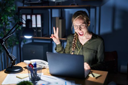 Photo for Young blonde woman working at the office at night smiling with happy face winking at the camera doing victory sign. number two. - Royalty Free Image