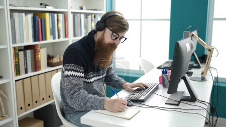 Foto de Young redhead man student using computer and headphones writing on notebook at library university - Imagen libre de derechos