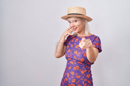 Foto de Mujer caucásica joven con vestido de flores y sombrero de verano sonriente haciendo hablar por teléfono gesto y señalando a usted. Llámame.. - Imagen libre de derechos