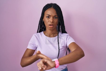 Photo for African american woman with braids standing over pink background in hurry pointing to watch time, impatience, upset and angry for deadline delay - Royalty Free Image