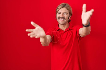 Téléchargez les photos : Homme caucasien avec moustache debout sur fond rouge regardant la caméra souriant à bras ouverts pour étreindre. expression joyeuse embrassant le bonheur. - en image libre de droit
