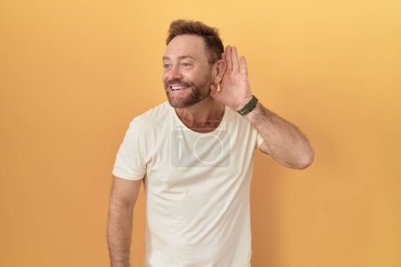 Photo for Middle age man with beard standing over yellow background smiling with hand over ear listening an hearing to rumor or gossip. deafness concept. - Royalty Free Image
