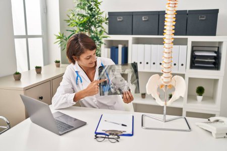 Foto de Joven hermosa mujer hispana médico señalando con el dedo a la columna vertebral de rayos X en la clínica - Imagen libre de derechos