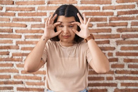 Photo for Young hispanic woman standing over bricks wall trying to open eyes with fingers, sleepy and tired for morning fatigue - Royalty Free Image