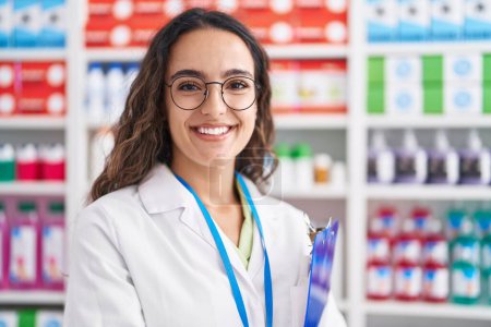 Téléchargez les photos : Jeune belle hispanique femme pharmacien souriant confiant tenant presse-papiers à la pharmacie - en image libre de droit