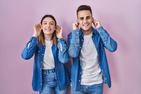 Photo for Young hispanic couple standing over pink background smiling pulling ears with fingers, funny gesture. audition problem - Royalty Free Image