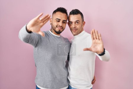 Foto de Pareja homosexual de pie sobre fondo rosa haciendo marco usando manos palmas y dedos, perspectiva de la cámara - Imagen libre de derechos