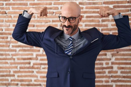Téléchargez les photos : Homme chauve avec barbe portant des vêtements de travail et des lunettes montrant les muscles des bras souriant fier. concept de fitness. - en image libre de droit