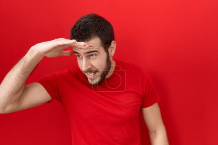Photo for Young hispanic man wearing casual red t shirt very happy and smiling looking far away with hand over head. searching concept. - Royalty Free Image