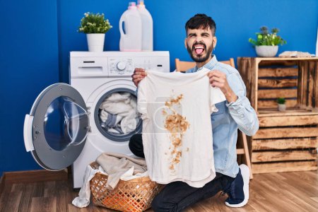 Foto de Joven hombre hispano con barba sosteniendo camiseta blanca limpia y camiseta con mancha sucia sacando la lengua feliz con expresión divertida. - Imagen libre de derechos