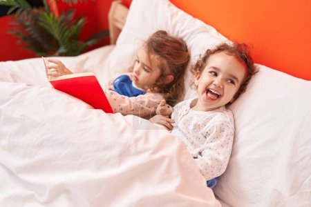 Photo for Adorable girls reading book sitting on bed at bedroom - Royalty Free Image