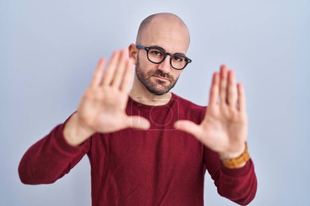Foto de Joven hombre calvo con barba de pie sobre fondo blanco con gafas haciendo montura usando las manos palmas y los dedos, perspectiva de la cámara - Imagen libre de derechos