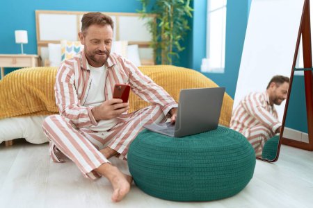 Foto de Hombre de mediana edad con teléfono inteligente y portátil sentado en el suelo en el dormitorio - Imagen libre de derechos