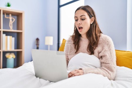Téléchargez les photos : Jeune femme hispanique utilisant un ordinateur portable sur le lit effrayé et étonné avec la bouche ouverte pour la surprise, visage incrédule - en image libre de droit