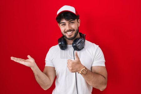 Téléchargez les photos : Homme hispanique avec barbe coiffé d'un chapeau de joueur et d'écouteurs montrant la main de la paume et faisant un geste correct avec les pouces levés, souriant heureux et joyeux - en image libre de droit