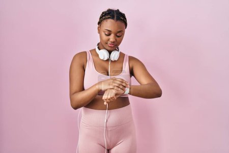 Téléchargez les photos : Femme afro-américaine avec des tresses portant des vêtements de sport et écouteurs vérifiant l'heure sur la montre-bracelet, détendu et confiant - en image libre de droit