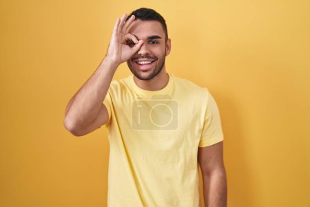 Photo for Young hispanic man standing over yellow background doing ok gesture with hand smiling, eye looking through fingers with happy face. - Royalty Free Image