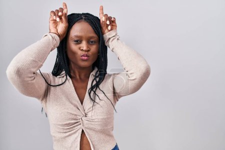 Téléchargez les photos : Femme africaine avec des tresses debout sur fond blanc faisant un geste drôle avec doigt sur tête comme des cornes de taureau - en image libre de droit