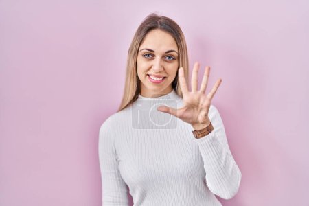 Foto de Mujer rubia joven con suéter blanco sobre fondo rosa mostrando y apuntando hacia arriba con los dedos número cinco mientras sonríe confiado y feliz. - Imagen libre de derechos